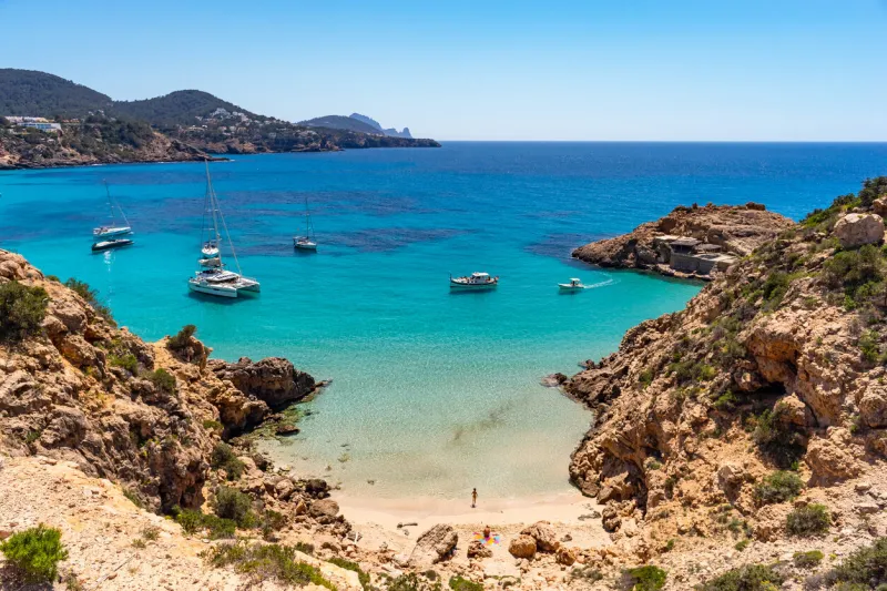 wide-angle view of the cove of platja des pujolets, near cala tarida, on the western coast of ibiza, fishermen's huts, warm light, a few boats rocking on transparent waters, the gentle splashing of the waves on the shore developed from raw