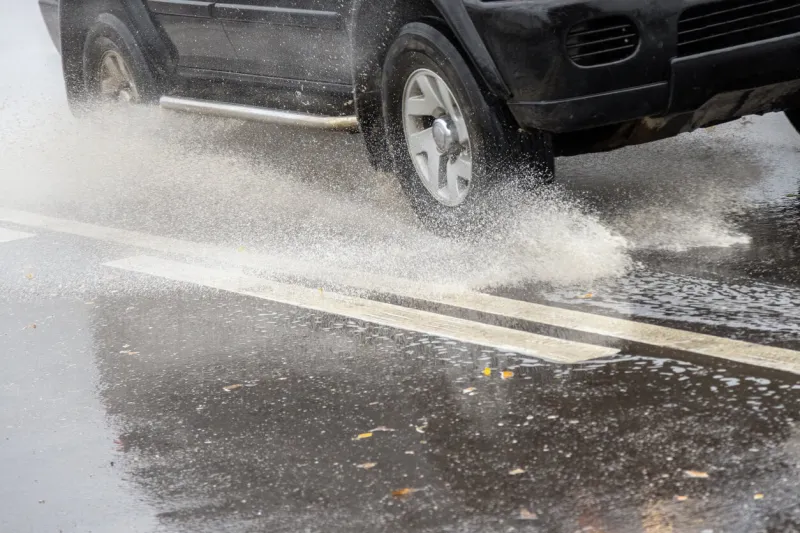 black suv rushes into shallow puddle on wet autumnal asphalt road at cloudy day light with water splashes