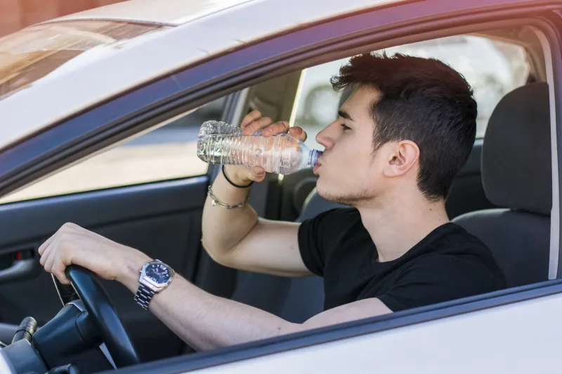 handsome young man or teenager driving car and drinking water from plastic bottle
