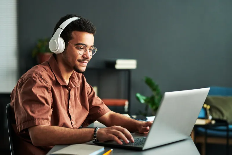 young smiling man in headphones typing on laptop keyboard while sitting by workplace and taking part in online webinar or lesson