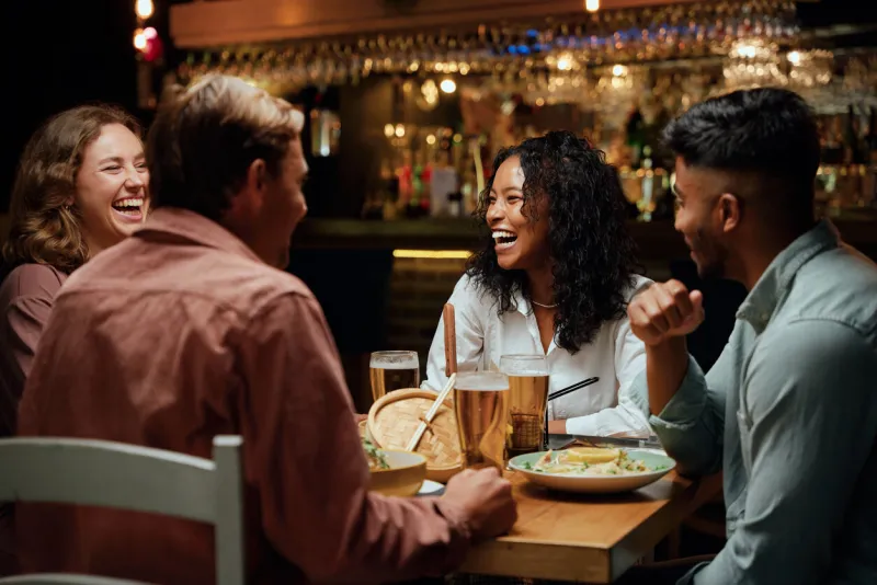 young multiracial group of friends wearing casual clothing laughing around table with food and drinks