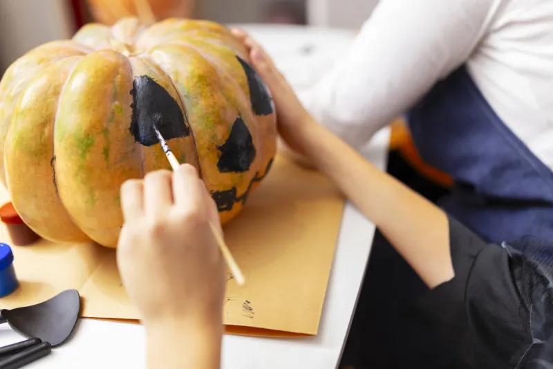 adult hand guiding child's hand in painting black face on orange pumpkin close-up photography for decoration and seasonal design