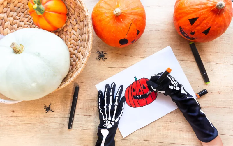 on a wooden desk surrounded by pumpkins and spiders, hands in black gloves with bones are drawing a sketch with markers a laughing pumpkin, top view preparation for the holiday halloween