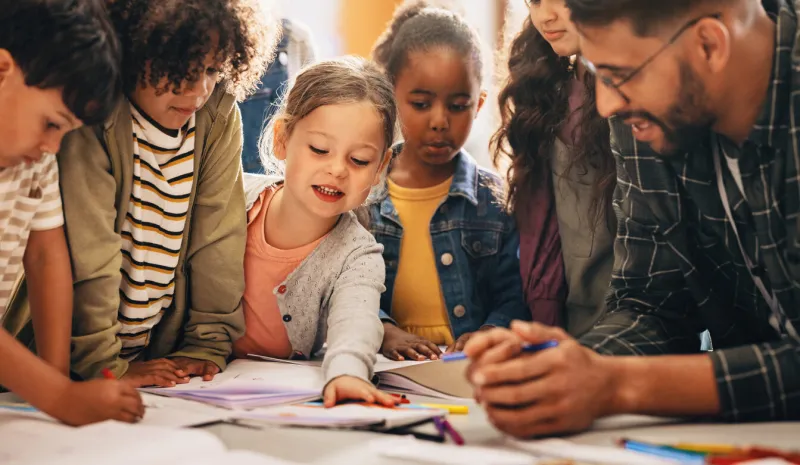 students doing a creativity project with their teacher in a classroom group of primary school children learning how to draw and colour with the help of their educator
