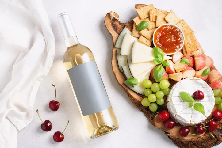 bottle of white wine mockup with blank label, cheese board and fruits on light gray background, top view