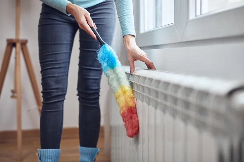 woman with a dust stick cleaning central heating gas radiator at home