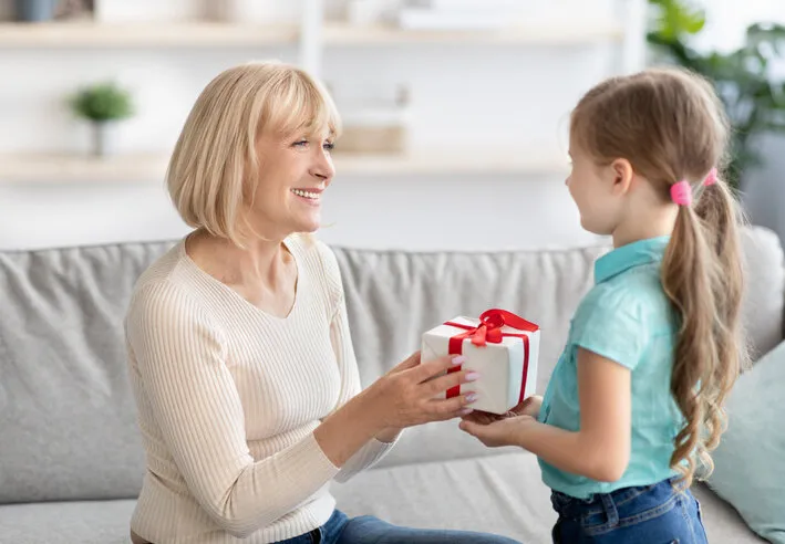 celebrating concept portrait of cute little girl making surprise for her grandma holding and giving present box, greeting woman with holiday granddaughter congratulating smiling mature lady