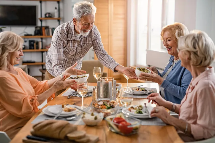happy mature man serving food while having lunch with female friends at home