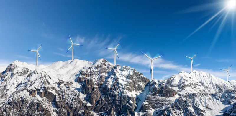 group of white and blue wind turbines on the top of snow capped mountains against a clear blue sky with clouds and sunbeams