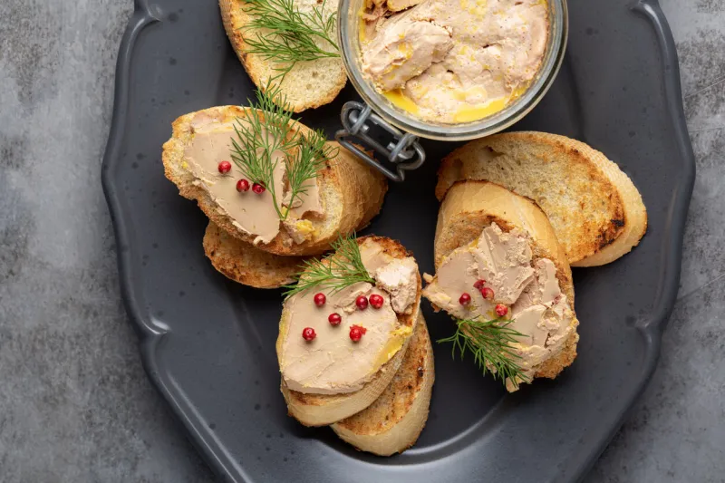 bread or baguette toast with foie gras pate, directly above a specialty food product made of the liver of a duck or goose, in a glass jar decorated with red pepper and dill