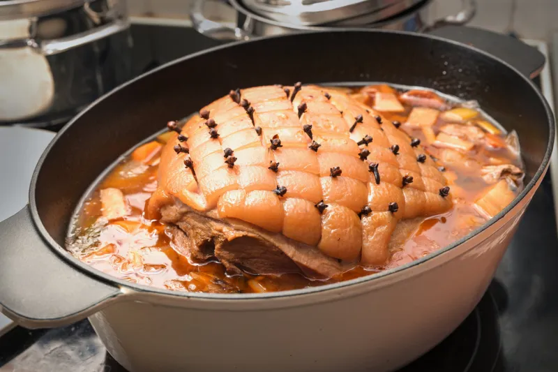 cooking a festive meal, roast pork with rind in diamond shapes and larded with cloves laid in a casserole with vegetables and broth ready for the oven, selected focus, narrow depth of field