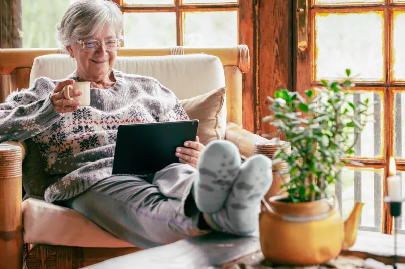 old senior woman sitting at home on armchair using digital tablet wearing a warm sweater and eyeglasses comfortable living room, wooden rustic windows