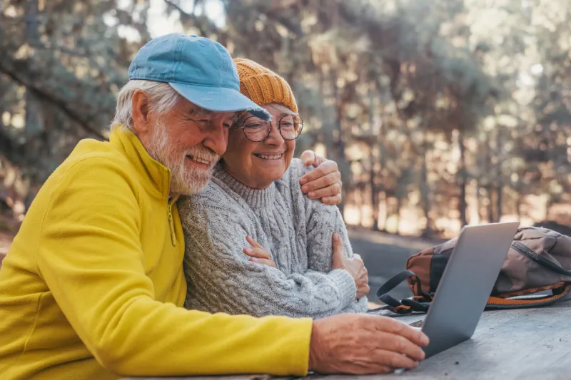 head shot portrait close up of cute couple of old middle age people using computer pc outdoors sitting at a wooden table in the forest of mountain in nature with trees around them
