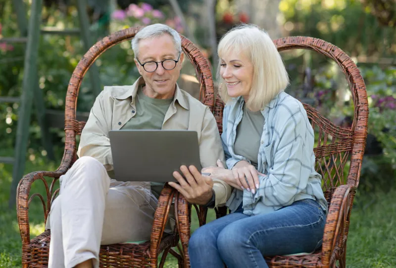modern senior spouses relaxing in wicker chairs in open air and using laptop computer, surfing internet or shopping online, enjoying warm spring evening outdoors, copy space