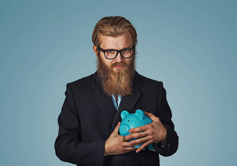 young greedy stingy bearded hipster businessman man holding piggy bank isolated on blue studio wall background negative face expression human emotion body language reaction attitude horizontal