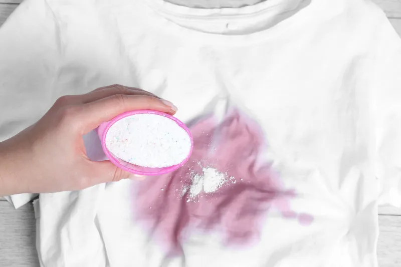 woman adding powdered detergent onto white t-shirt with stain on wooden surface, top view hand washing laundry