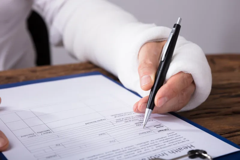 close-up of a businessperson with broken arm filling health insurance claim form on wooden desk