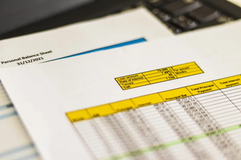 close up shot of a print outs of excel table of a bank loan amortization table, personal balance sheet and laptop banking