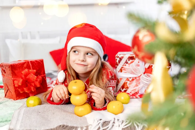 a girl child on the bed at home playing with tangerines building a tangerine snowman and waiting for the new year or christmas in a red santa claus hat