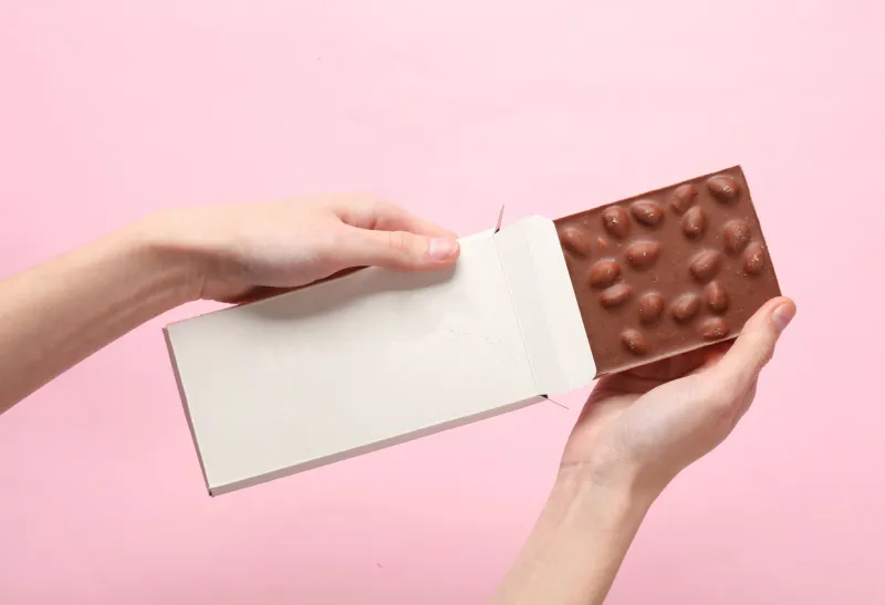 hands holding mockup of white cardboard box with chocolate bar on pink background