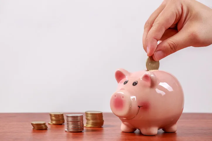 close-up of hand putting coin in piggy bank on table against white background