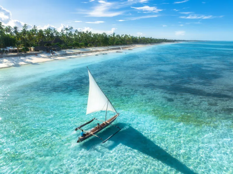 aerial view of the sailboat on blue sea, empty white sandy beach at sunset summer vacation in zanzibar tropical landscape with boat, ocean with clear water, green palms, sky top drone view exotic