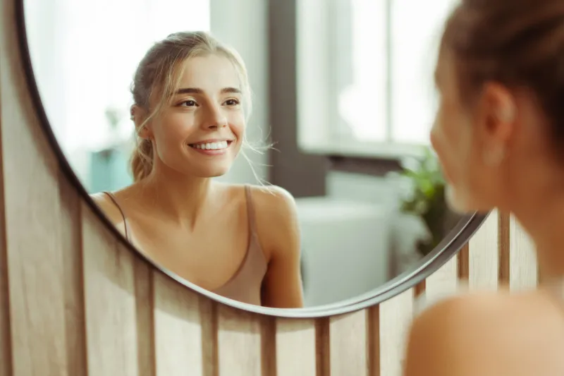 smiling, beautiful, confident woman with make up wearing tank top, looking in mirror, standing in bathroom at home skin care concept, morning routine