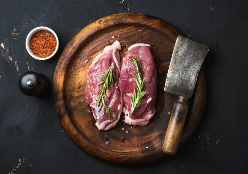 raw uncooked poultry meat cut duck breast with rosemary, spices and butcher cleaver on dark wooden tray over black wooden background, top view