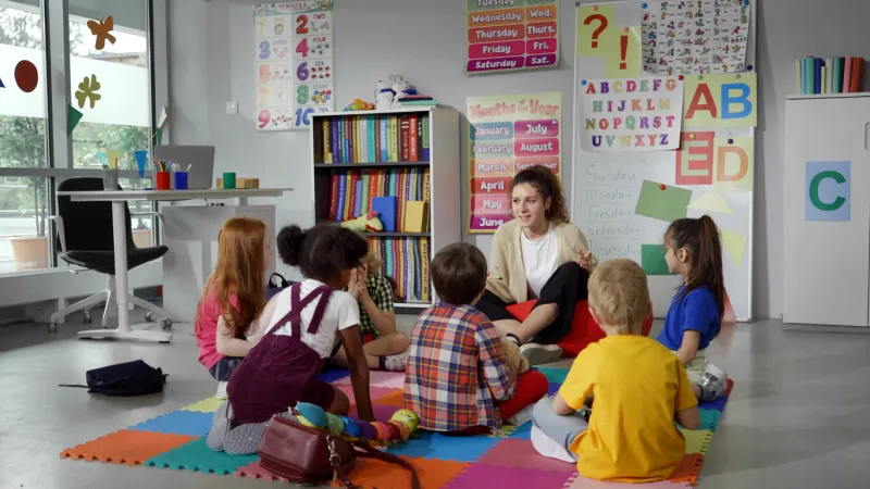 small nursery school children with teacher sitting on floor having lesson young woman teach preschool kids in classroom sitting on floor together