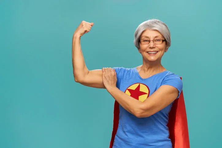 joyful beautiful senior woman in superhero costume posing on turquoise background