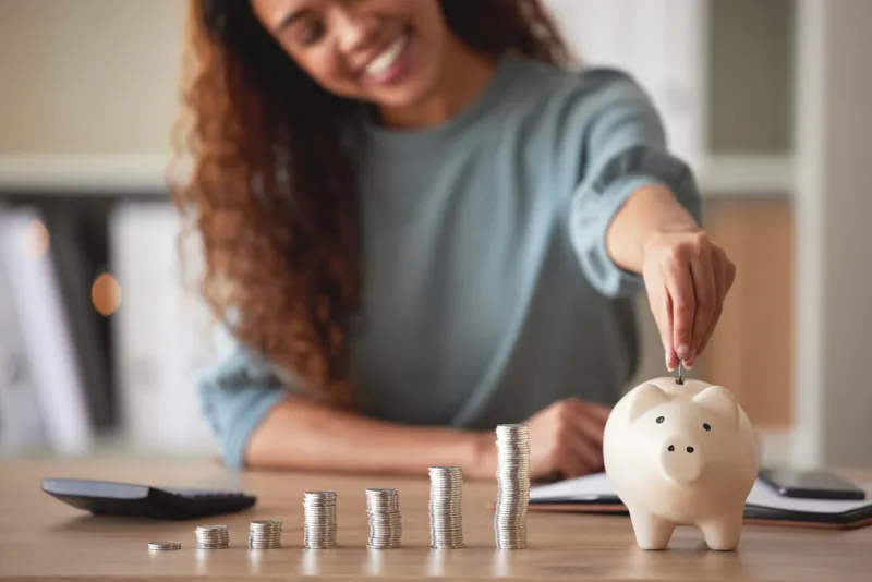 young african american woman money putting coins into a piggybank at home mixed race person counting coins while financial planning in her living room saving, investing and thinking about the future