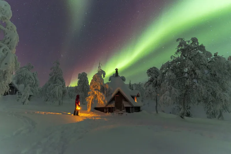 hiker with lantern beside a frozen hut in the snowy forest under the northern lights, finland