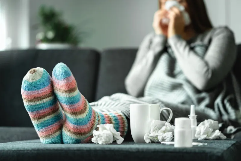 sick woman with flu, cold, fever and cough sitting on couch at home ill person blowing nose and sneezing with tissue and handkerchief woolen socks and medicine infection in winter resting on sofa
