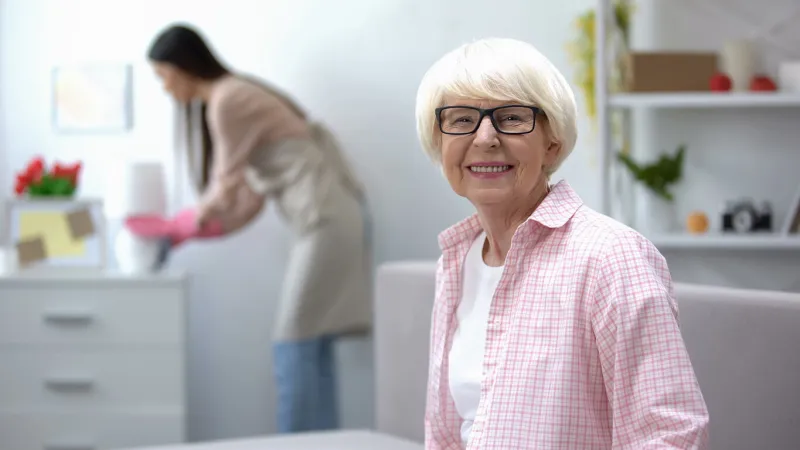 smiling aged woman looking at camera, housekeeper wiping dust in room, cleaning