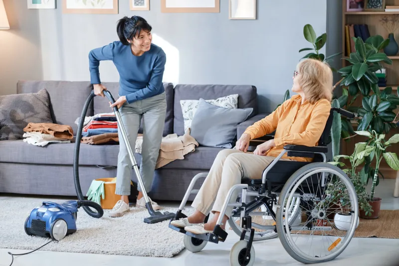 housemaid using vacuum cleaner to clean the carpet in the room, she helping senior woman who using wheelchair