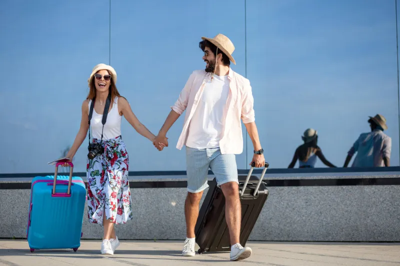 happy young couple walking in front of an airport terminal building, pulling suitcases