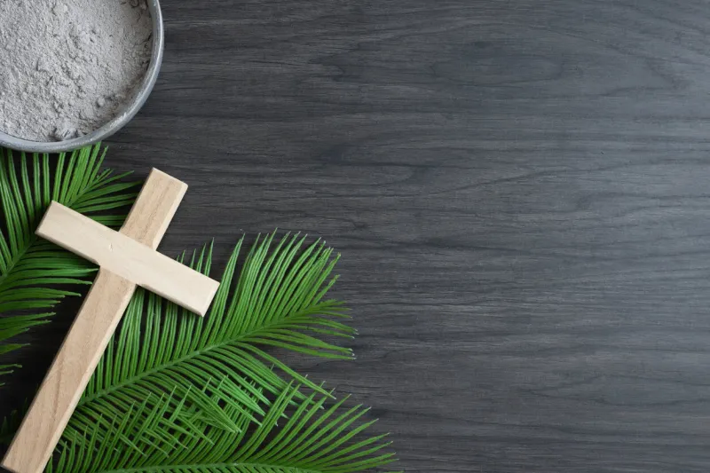 christian lenten border of a bowl of ashes, palm leaves and simple wood cross on a dark wood background with copy space