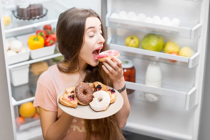young woman in the sleepwear eating sweet donuts near the refrigerator