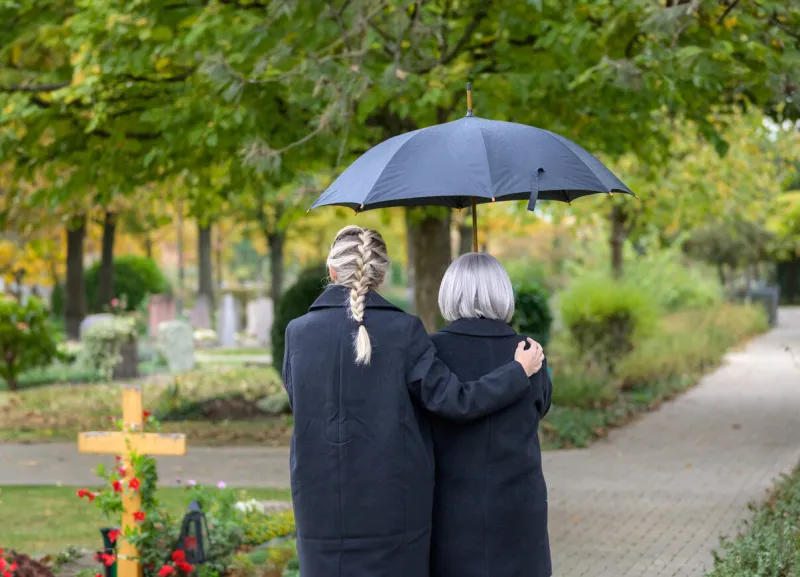 rear view of daughter holding her mother in her arms on the way to the man's grave in the cemetery on a cloudy cold day