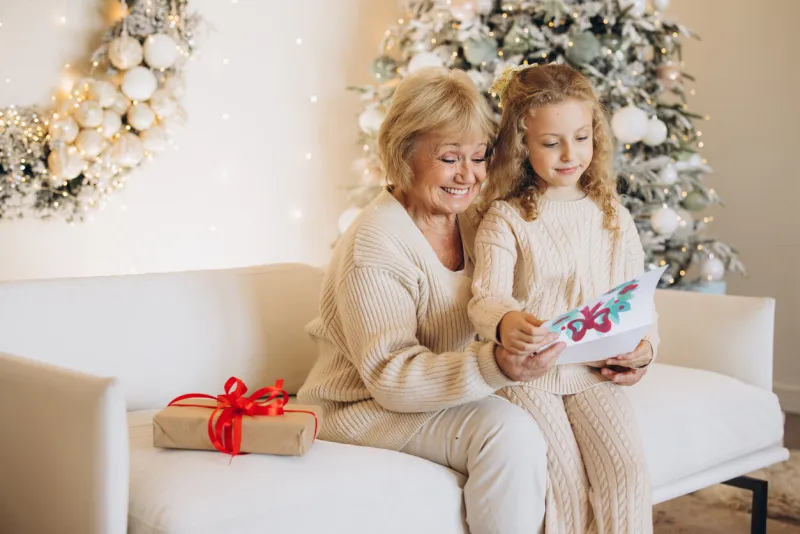 an elderly woman and her young granddaughter enjoy reading a christmas card together on a cozy couch, surrounded by festive holiday decorations and a beautifully adorned tree