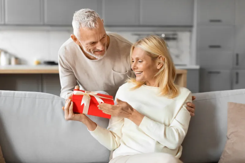 loving senior man greeting his excited attractive blonde wife with valentines day, husband hugging spouse from behind and giving her red gift box birthday party, anniversary celebration