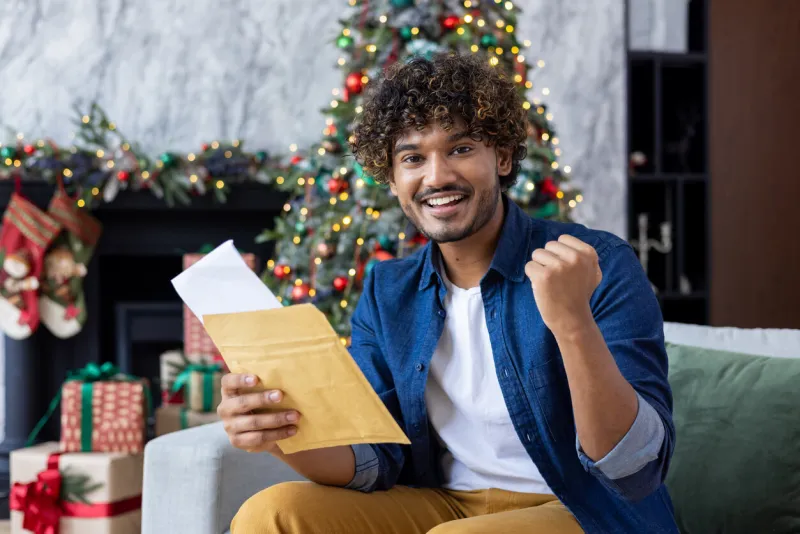 portrait of indian young man rejoicing in success and achievement, showing yes hand gesture sitting at home near the christmas tree, holding a letter in his hands and smiling at the camera