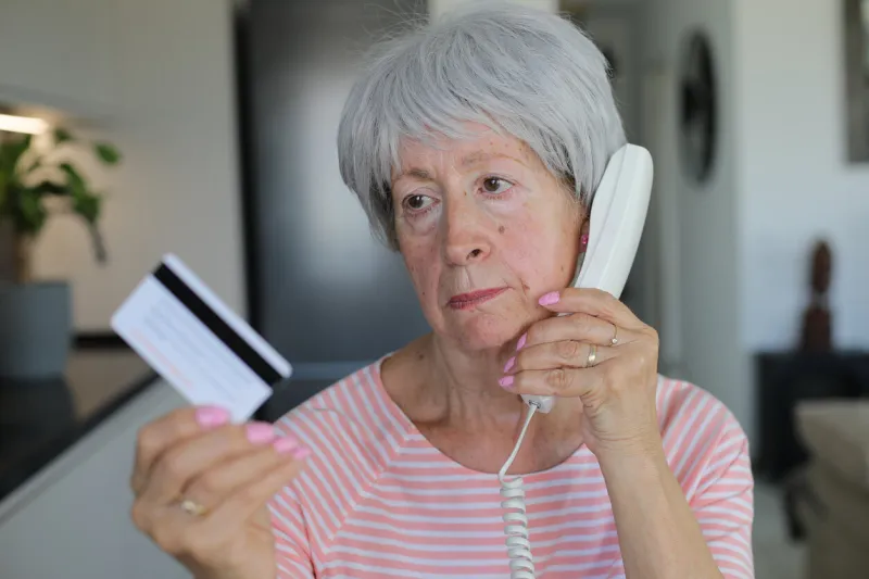 a pretty and skeptical looking senior caucasian woman in her late sixties is holding a credit card during phone call on her land line