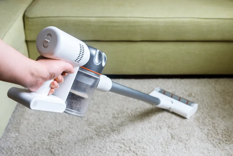 wireless vacuum cleaner used on carpet in room housework with new upright hoover person holds modern white vacuum cleaner by sofa home cleaning, care and technology concept