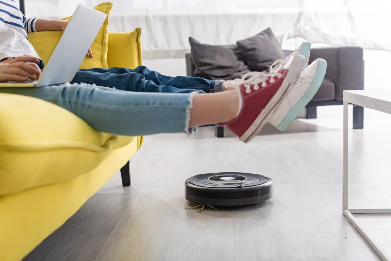 cropped view of kid with laptop near mother with raised legs on sofa and robotic vacuum cleaner on floor in living room