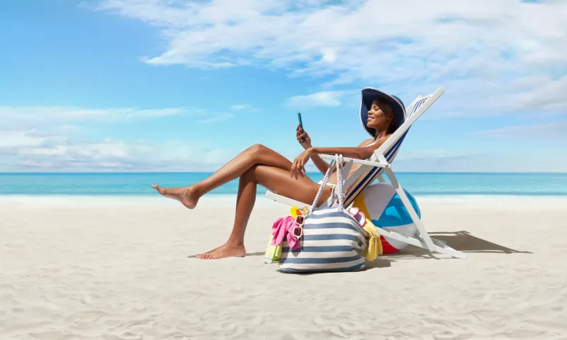 happy woman at the beach on beach deck chair, sunbathing, uses mobile phone, in a sunny day with blue sky, concept a summer beach holiday, online shopping, booking travel, and resorts accommodations