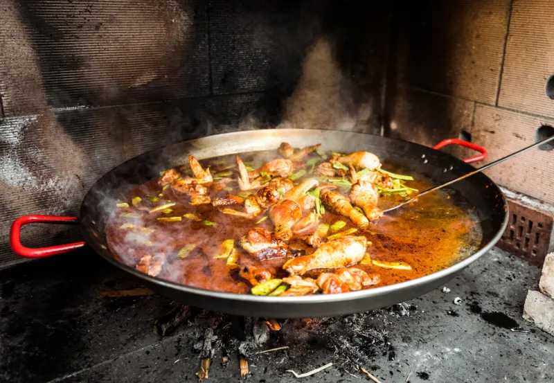 cooking and making a traditional spanish paella over open fire traditional way of preparing valencian paella with fire wood and flames in a big pan made from chicken and rabbit meat in fireplace