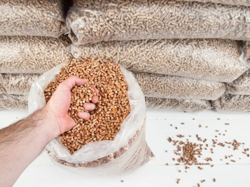 mans hand gripping woodpellets with contents of an open bag of wood pellets and a stack of bagged wood pellets behind