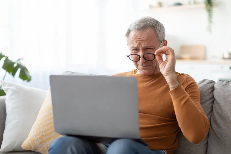 poor eyesight senior man squinting eyes using laptop wearing eyeglasses having problems with vision sitting on couch ophtalmic issue, bad sight in older age, macular degeneration concept