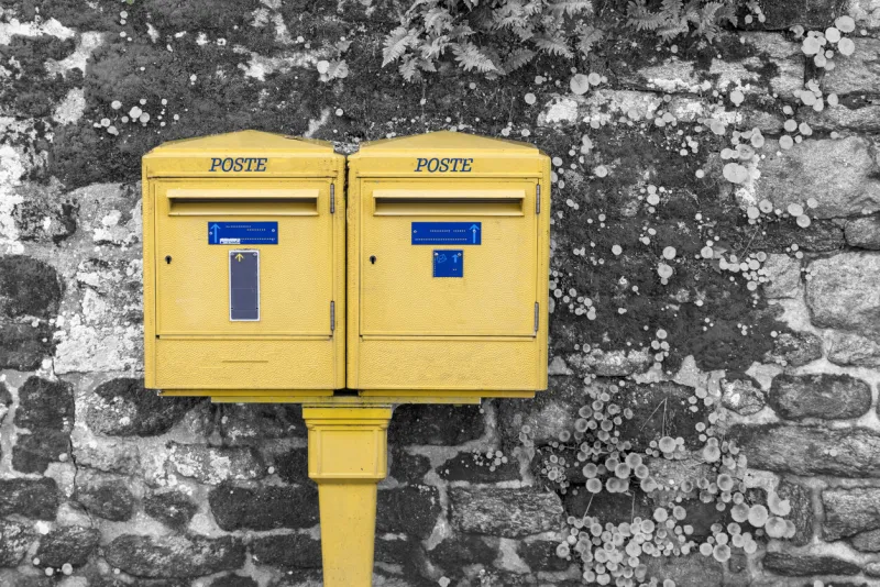 two french letterboxes, with the word poste written on them, against a stone wall covered with vegetation